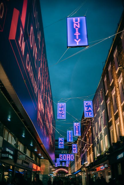 A view of Carnaby Street in Soho W1, featuring a large illuminated sign spelling 'SOHO' hanging above the pedestrianized street. The street is lined with historic brick buildings housing shops and caf