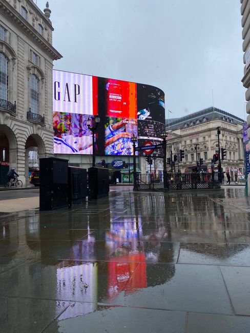 Nighttime view of Piccadilly Circus with illuminated digital advertising screens displaying brands such as Coca-Cola, Samsung, TDK, and Sanyo. The wet pavement reflects the vibrant colors and logos, i
